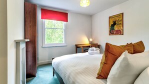 Une chambre avec un lit, une fenêtre avec un store rouge, une table avec une lampe et une affiche accrochée au mur.