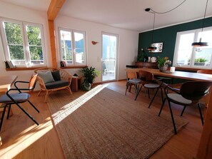 A spacious living area with a wooden floor, a large rug, and a dining table with chairs. There are large windows providing natural light and a view of greenery outside. - Woods: Accommodation **Haus im Glück** for large groups (St. Blasien)