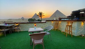 Un espace extérieur aménagé avec des sièges, offrant une vue sur les pyramides au coucher du soleil, comprenant des chaises en osier, une table agrémentée d’un vase de fleurs et un mur en pierre avec des tabourets de bar.