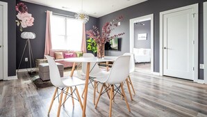 Un salon moderne avec un coin repas, comprenant une table et des chaises blanches, un canapé et une grande fenêtre avec des rideaux.