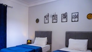 A hotel room with two beds, each with a blue bedspread and white pillows. There are four framed wall art pieces depicting cacti and a clock.