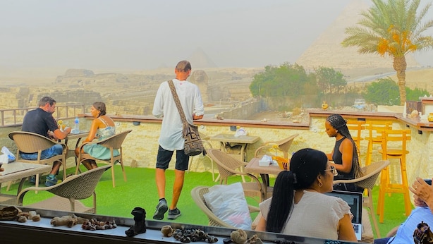 A terrace with wicker chairs and tables, overlooking a desert landscape with a pyramid in the distance.