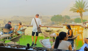 A terrace with wicker chairs and tables, overlooking a desert landscape with a pyramid in the distance. - Seven Pyramids (Gizah)