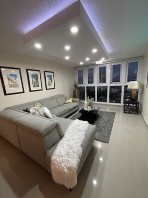 Ein modernes Wohnzimmer mit einem grauen Sofa, einem weißen, flauschigen Ottomanen und einem schwarzen Couchtisch. An der Wand hängen drei gerahmte Bilder, ein großes Fenster mit Blick auf die Stadt und eine Lampe auf einem Beistelltisch.