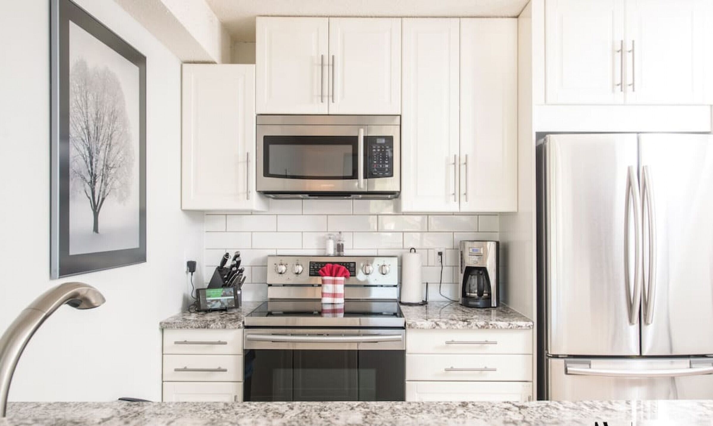 Una cocina moderna con gabinetes blancos, un refrigerador de acero inoxidable, un microondas, un horno y una cafetera.