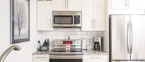 Una cocina moderna con gabinetes blancos, un refrigerador de acero inoxidable, un microondas, un horno y una cafetera.