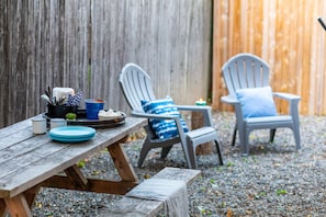 Une table de pique-nique en bois, avec des assiettes, des gobelets et une bougie. Deux chaises Adirondack avec des coussins bleus sont placées de chaque côté de la table.