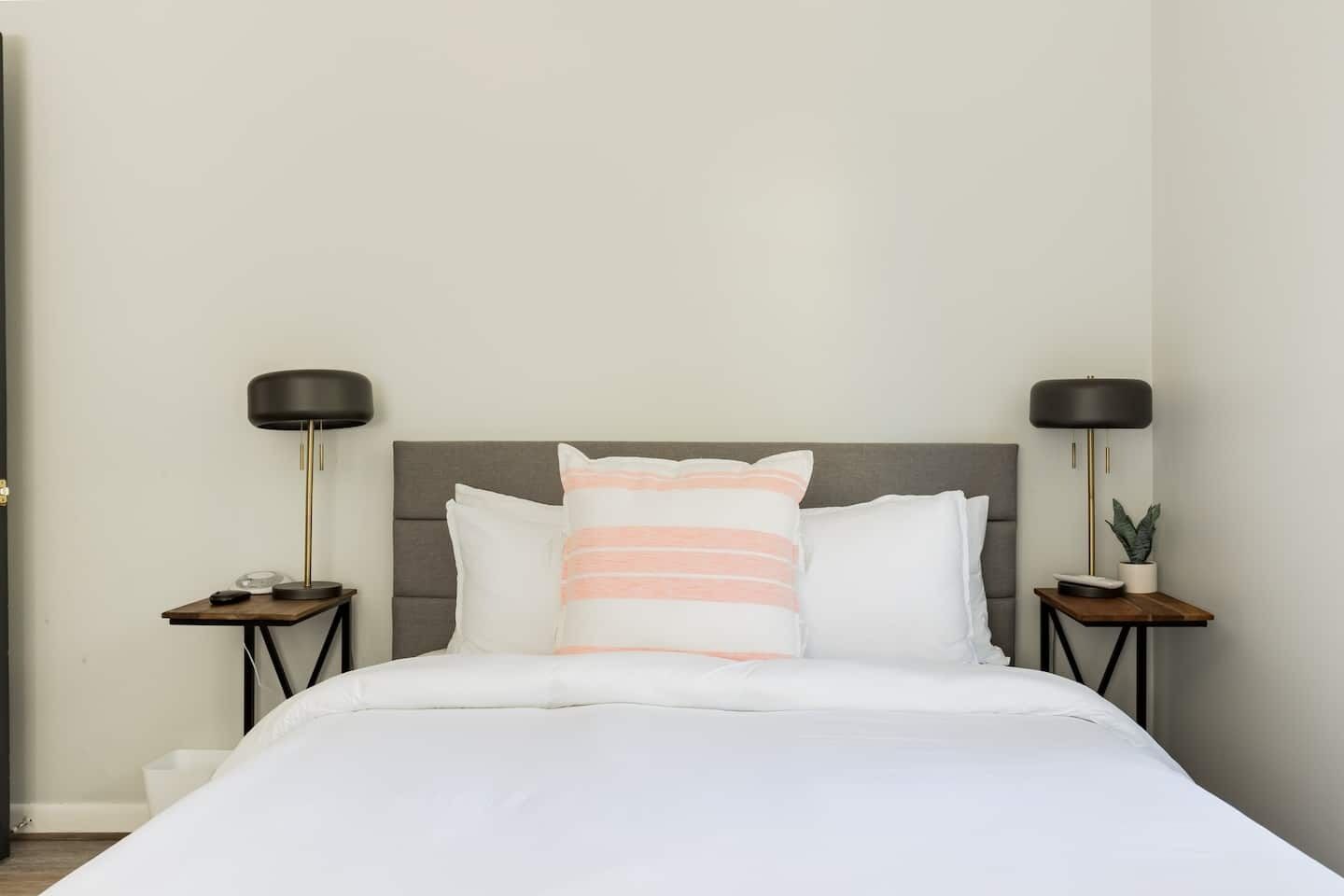 A neatly made bed with white bedding and a single striped pillow. There are two bedside tables, each with a black lamp and a small potted plant.