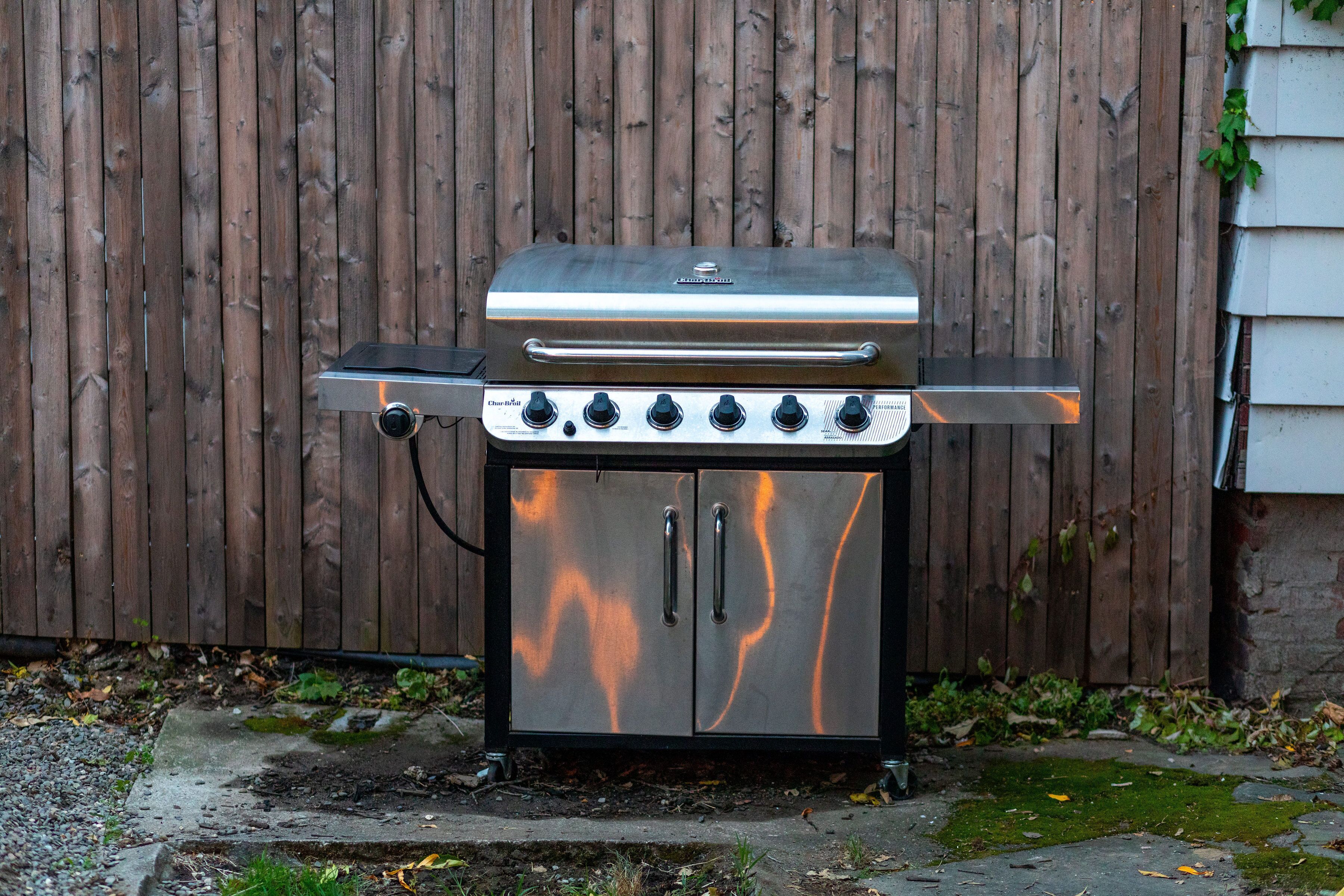 Een roestvrijstalen barbecue met vier branders en een extra brander, buiten geplaatst tegen een houten schutting.
