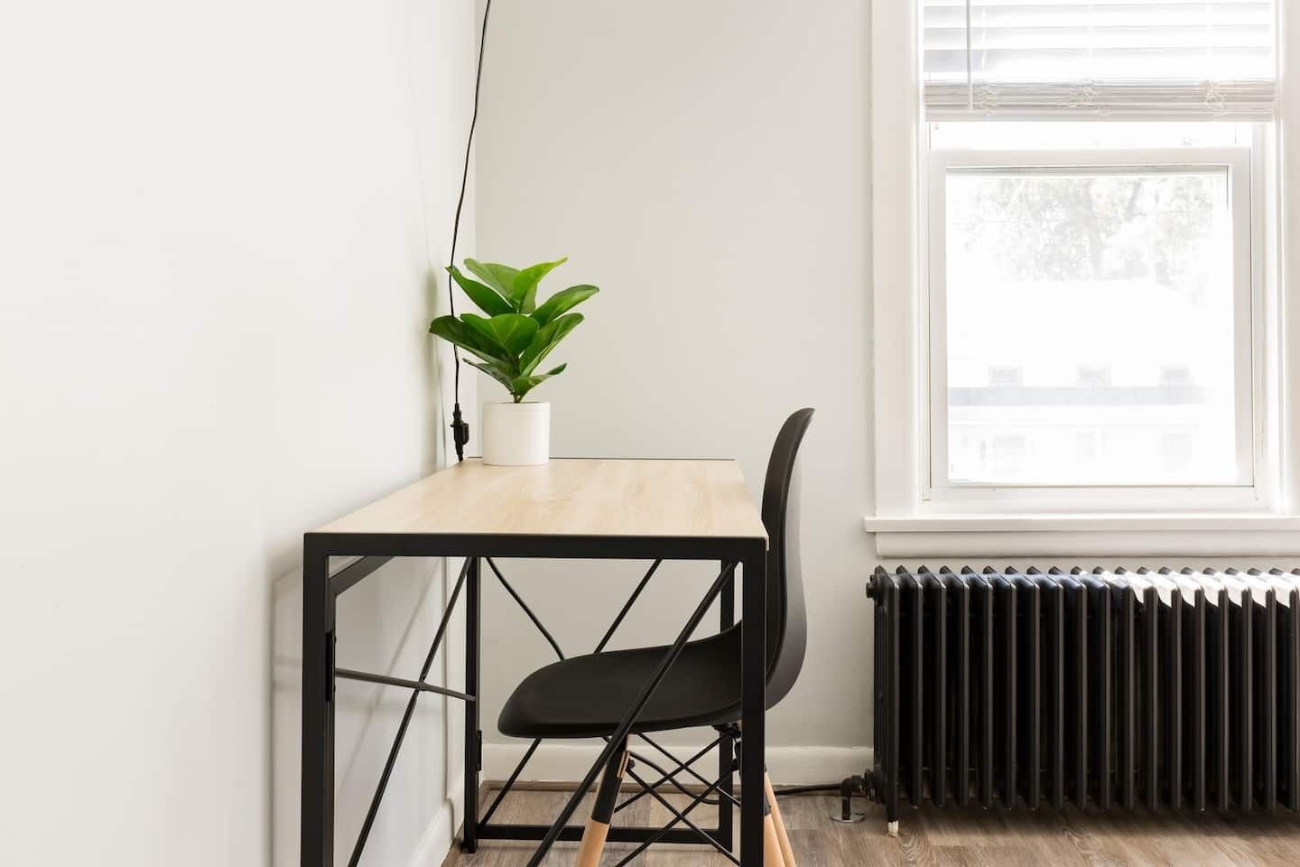 Et lille, moderne rum med et træbord og en sort stol, en potteplante på bordet, og en radiator under et vindue.