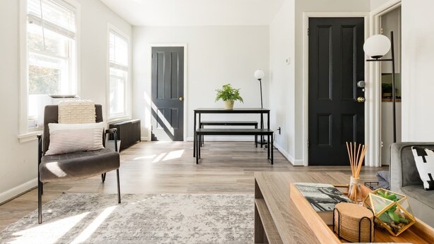 Een moderne woonkamer met een bank, een salontafel en een eettafel. Er zijn twee deuren, een zwarte en een witte, en een raam met zonwering.