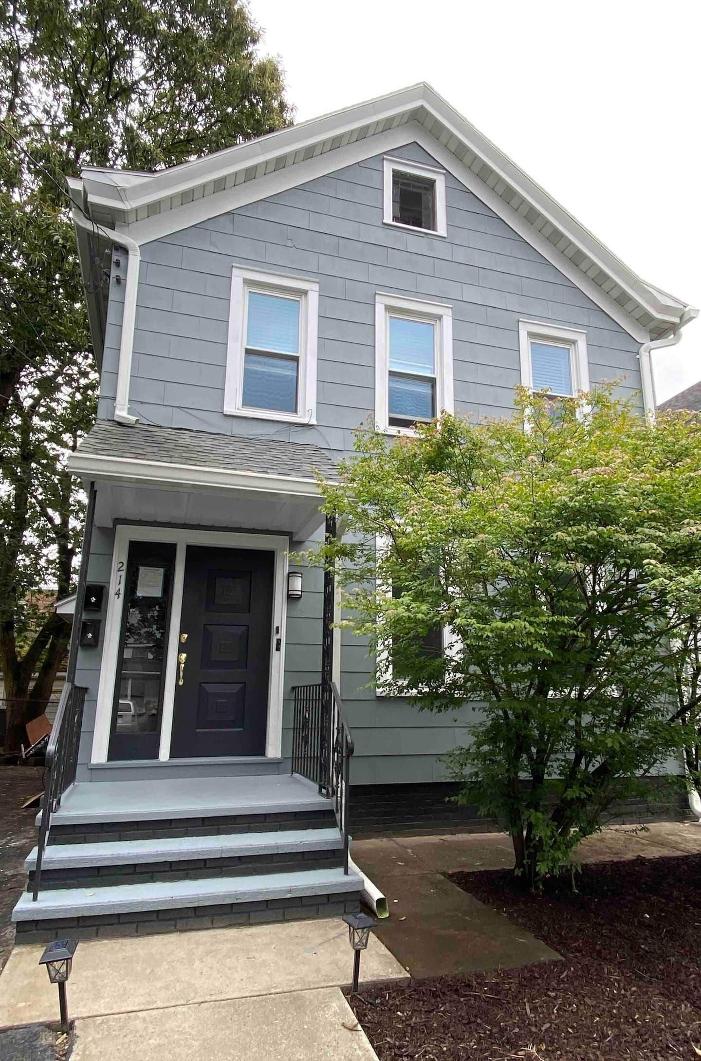A two-story house with a front porch, a small tree in front, and a mailbox.