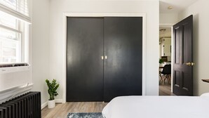 Een slaapkamer met een groot bed, een donkere dubbele deur, een radiator, een kleine potplant en uitzicht op de aangrenzende kamer.
