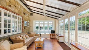 A sunlit room with stone walls, large windows, a sofa, a table, and a television. - Rookley Farm Lodge (Rookley)