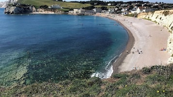 A coastal beach with clear water, a sandy shore, and buildings in the background.