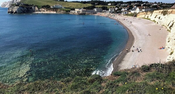 Una playa costera con agua cristalina, una orilla arenosa y edificios al fondo.