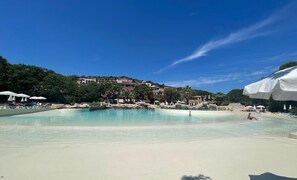 Zona de piscina amb aigua cristal·lina, platja de sorra i fons d'edificis i arbres.