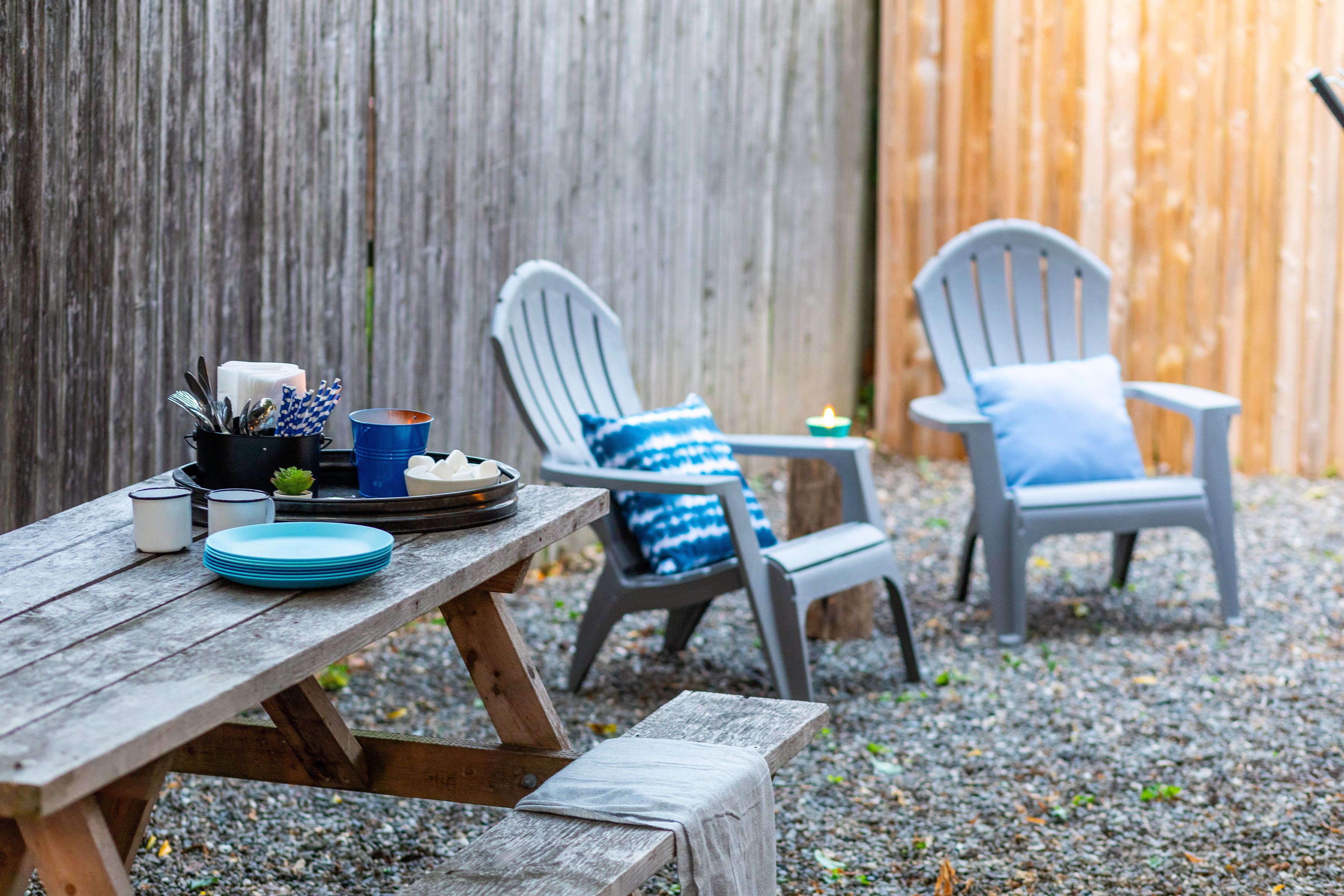 Una mesa de picnic de madera con platos, vasos y una vela. Dos sillas Adirondack con cojines azules están colocadas al lado de la mesa.