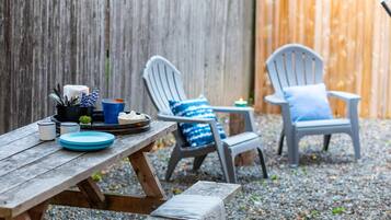 Una mesa de picnic de madera con platos, vasos y una vela. Dos sillas Adirondack con cojines azules están colocadas al lado de la mesa.