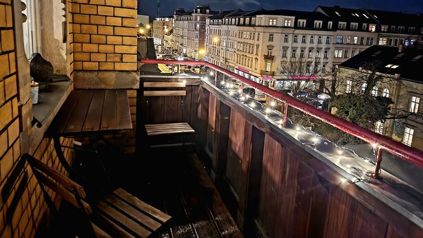 Un balcón con vista a una calle urbana de noche, que presenta una barandilla roja y bancos de madera.