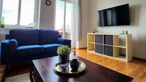 Una sala de estar con un sofá azul, una mesa de centro de madera con una planta y un jarrón, un televisor en la pared y una ventana con persianas.
