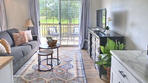 Un salon avec un canapé gris, une petite table ronde, un téléviseur posé sur un meuble et une vue sur une terrasse et la verdure à l’extérieur.