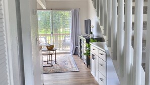 Un couloir avec un plancher en bois, un escalier avec des rampes blanches, un coin repas avec une table et des chaises, et une vue sur la verdure à l’extérieur.