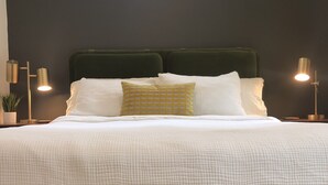 A neatly made bed with white linens and a green headboard, two bedside lamps, and a window with a view of greenery. -   Come enjoy this mountain retreat this Fall! (Saluda)