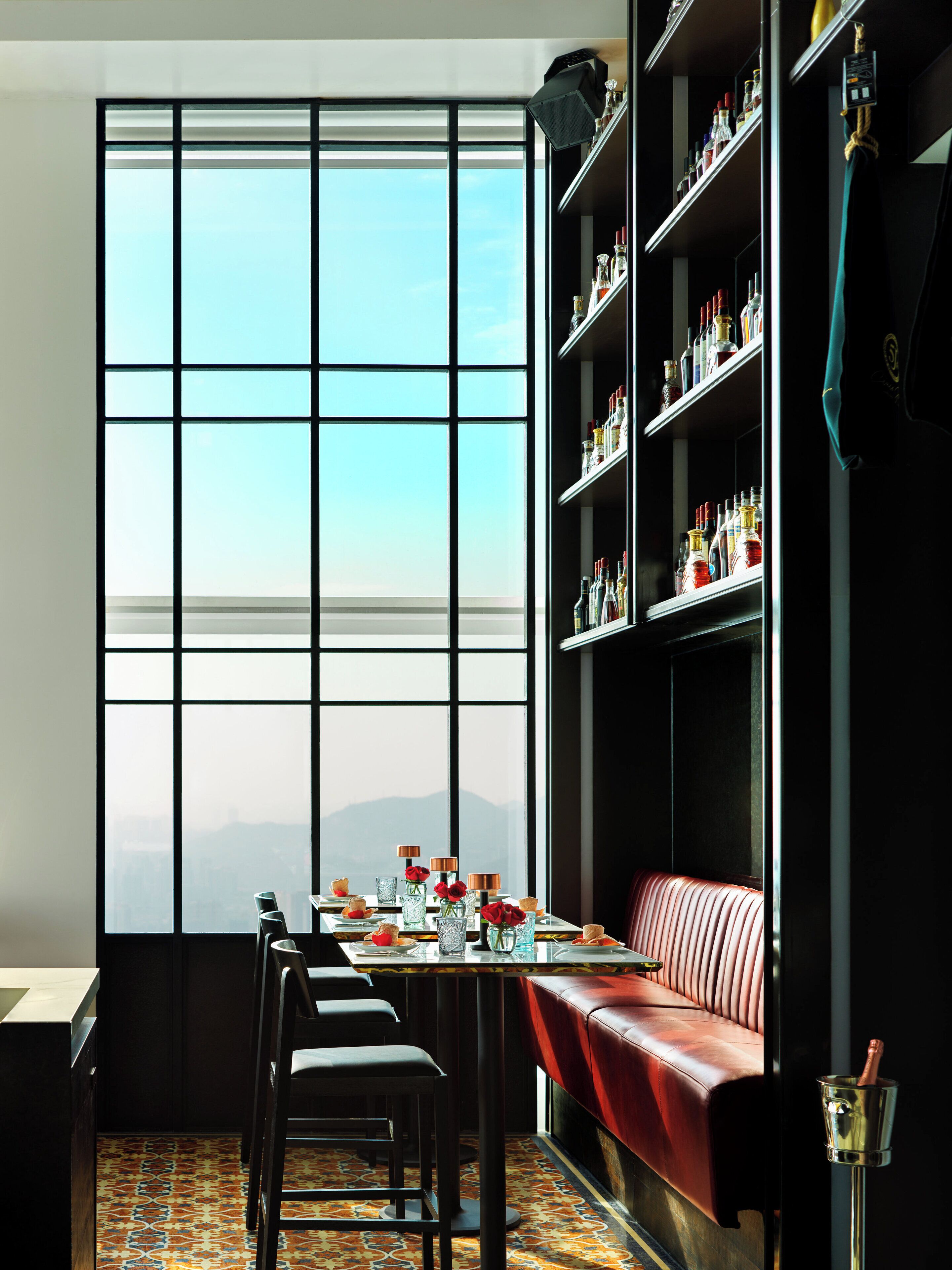 A modern interior with a large window, a bar area with shelves of bottles, and a red leather bench.