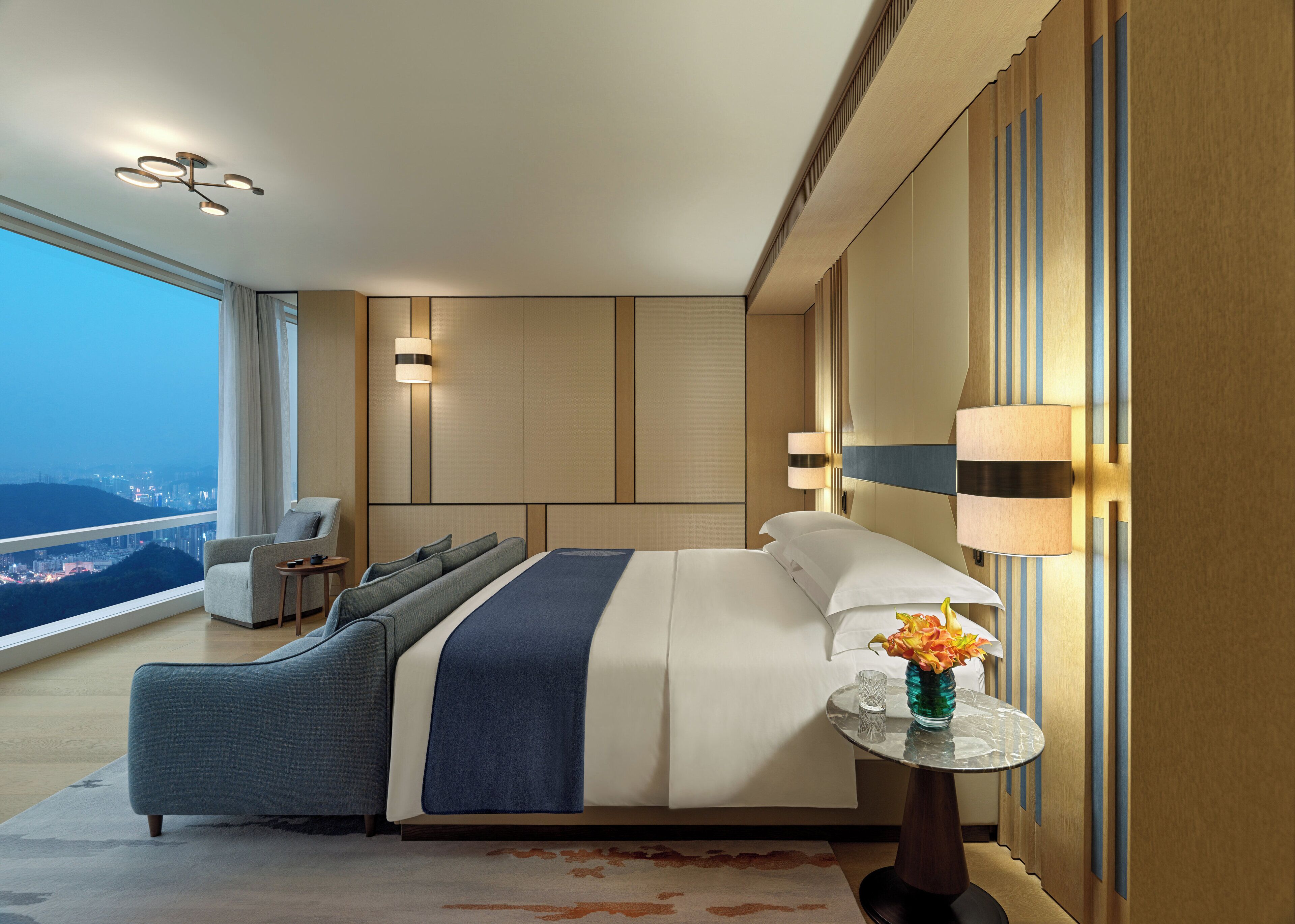 A modern hotel room with a large bed, a seating area with a chair and a small table, and a view of the cityscape.