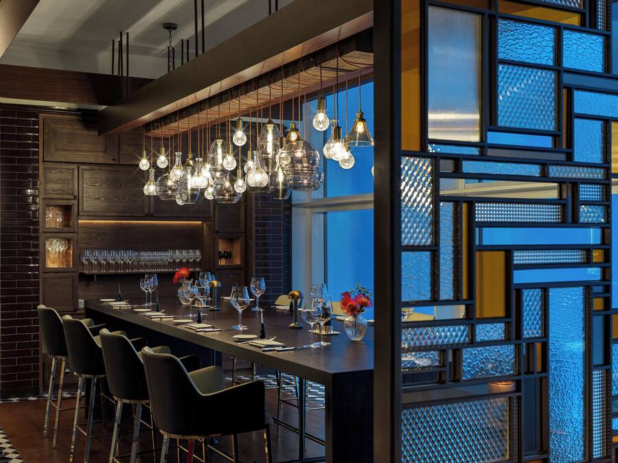 A modern bar area with a long counter, multiple hanging light fixtures, and a patterned glass partition.