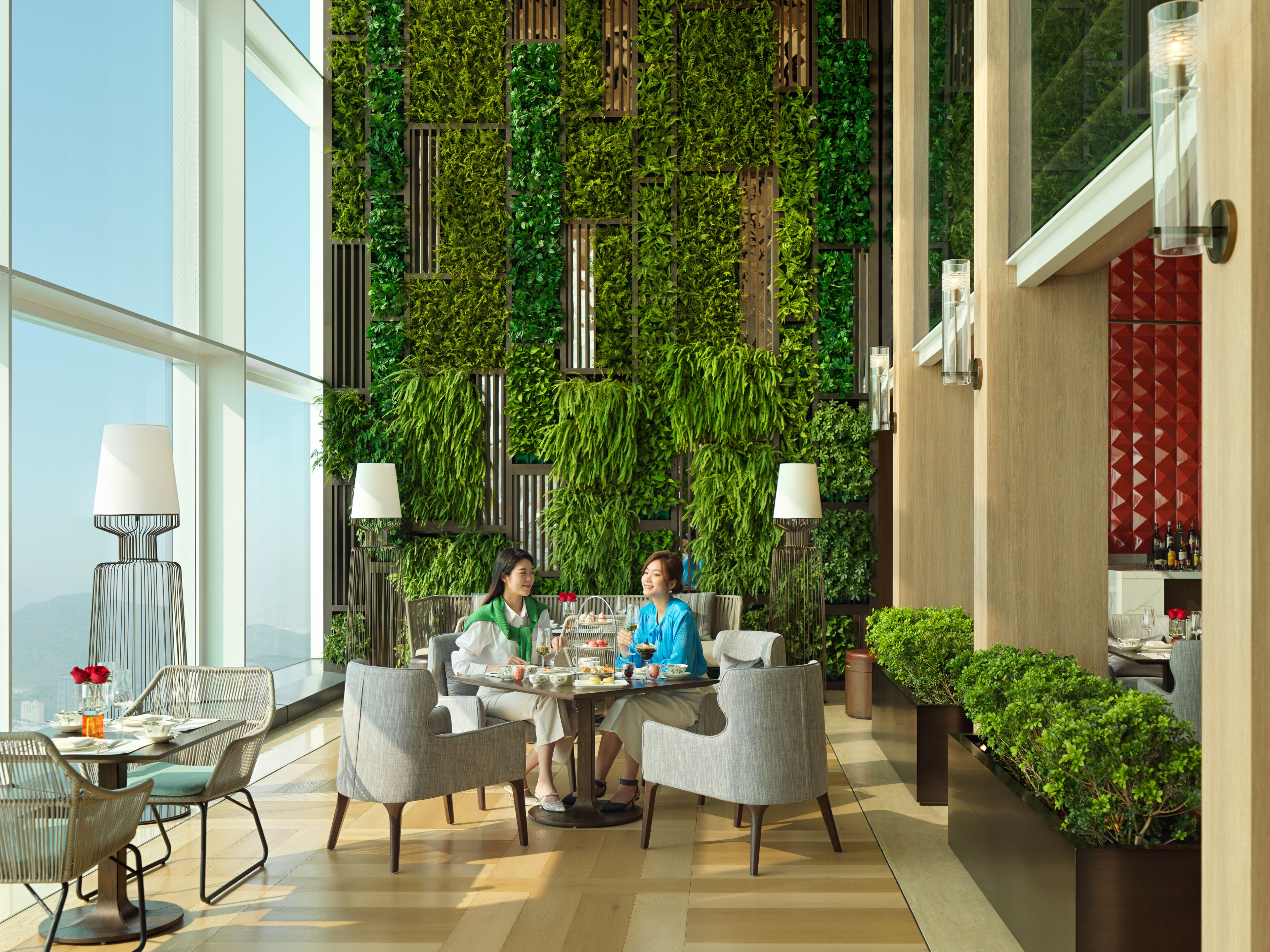 Un comedor moderno con techo alto, ventanas grandes y una pared cubierta de plantas verdes. Hay dos mesas de comedor con sillas, y una zona de bar con una partición de diseño rojo.
