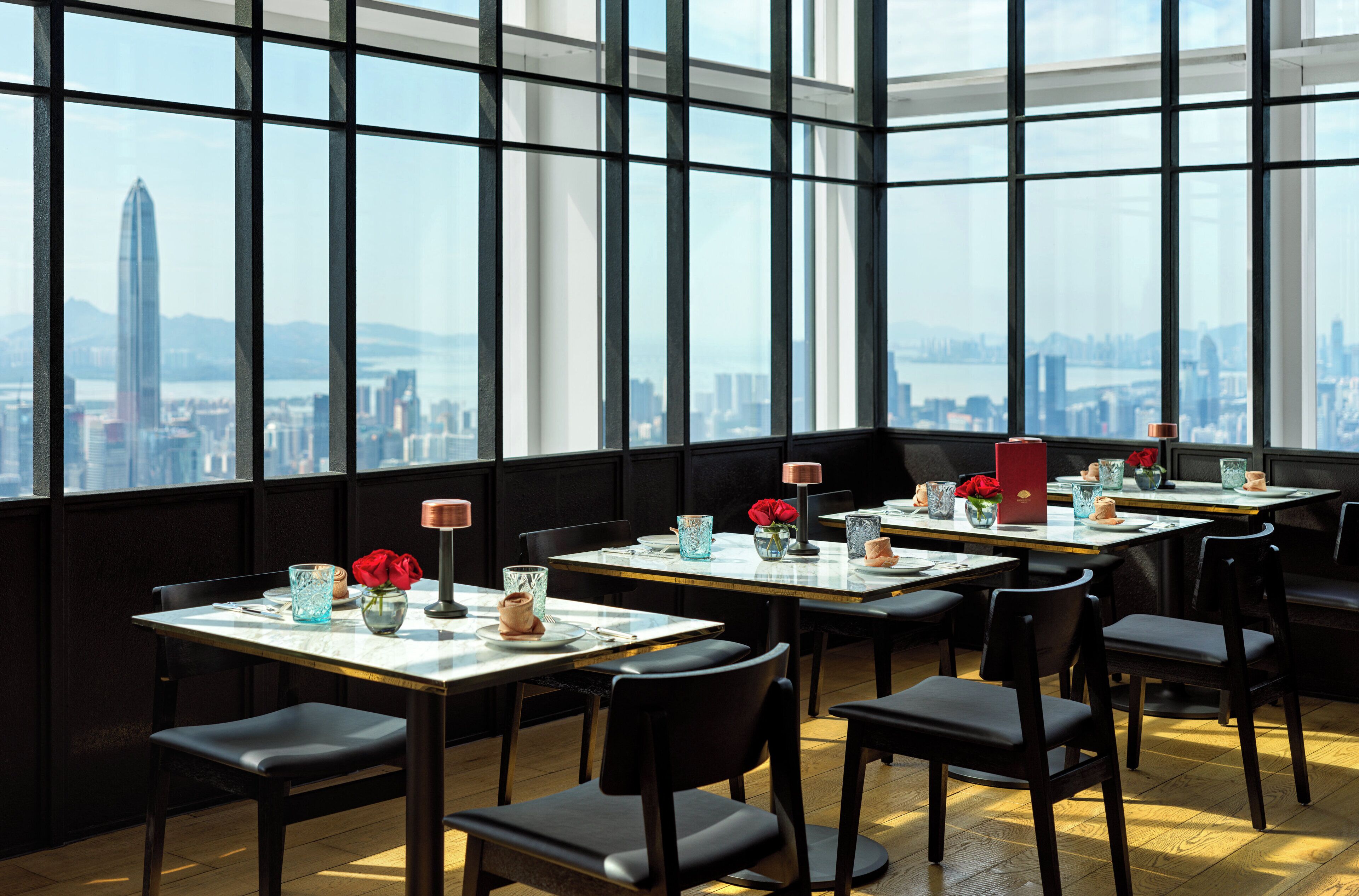 A restaurant with tables set for dining, large windows offering a city view, and modern interior design.