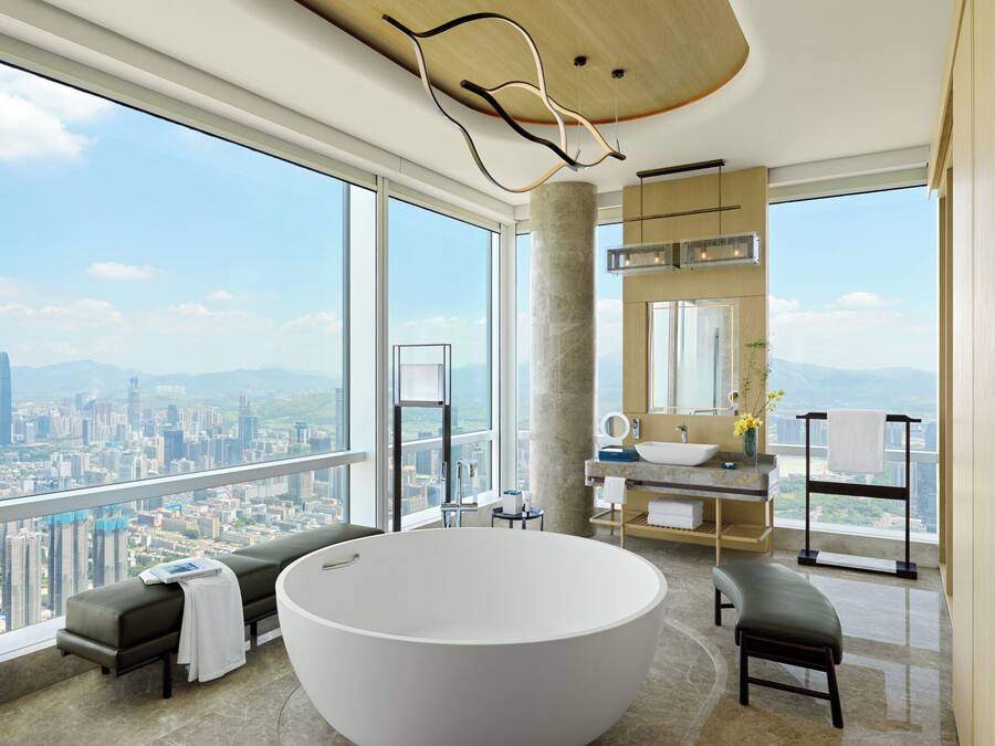 Un baño moderno con una bañera grande, un tocador con lavamanos y un espejo. Hay un banco y un perchero para toallas. El baño tiene ventanas amplias con vistas a la ciudad.