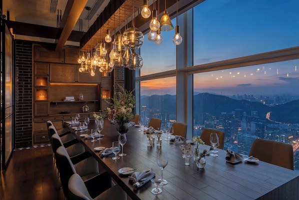A dining area with a long table set for a meal, large windows offering a city view, and hanging light fixtures.