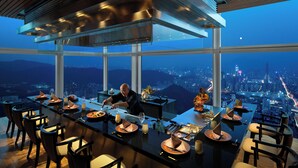 Zona de comedor en un edificio alto con vistas a la ciudad, cocina moderna y ventanas amplias.