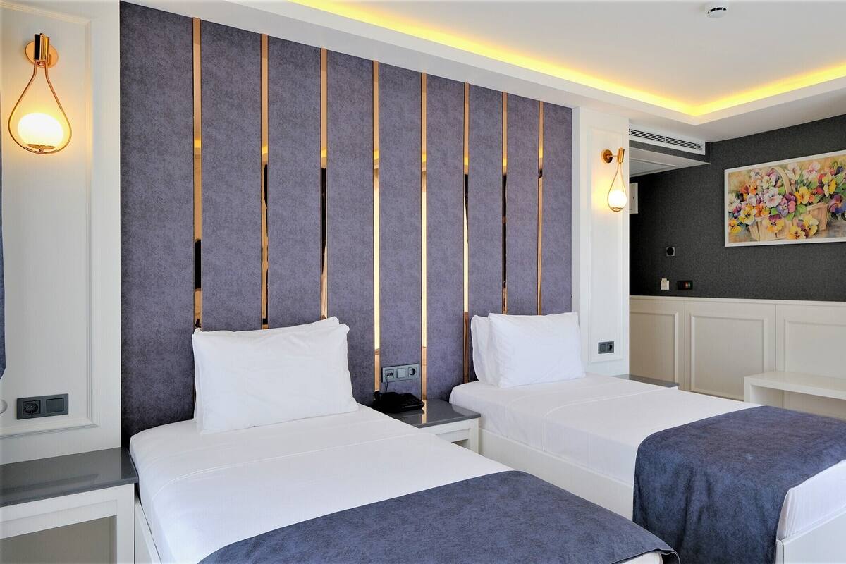 A modern hotel room with two beds, a gray accent wall, a painting, and a ceiling light fixture.