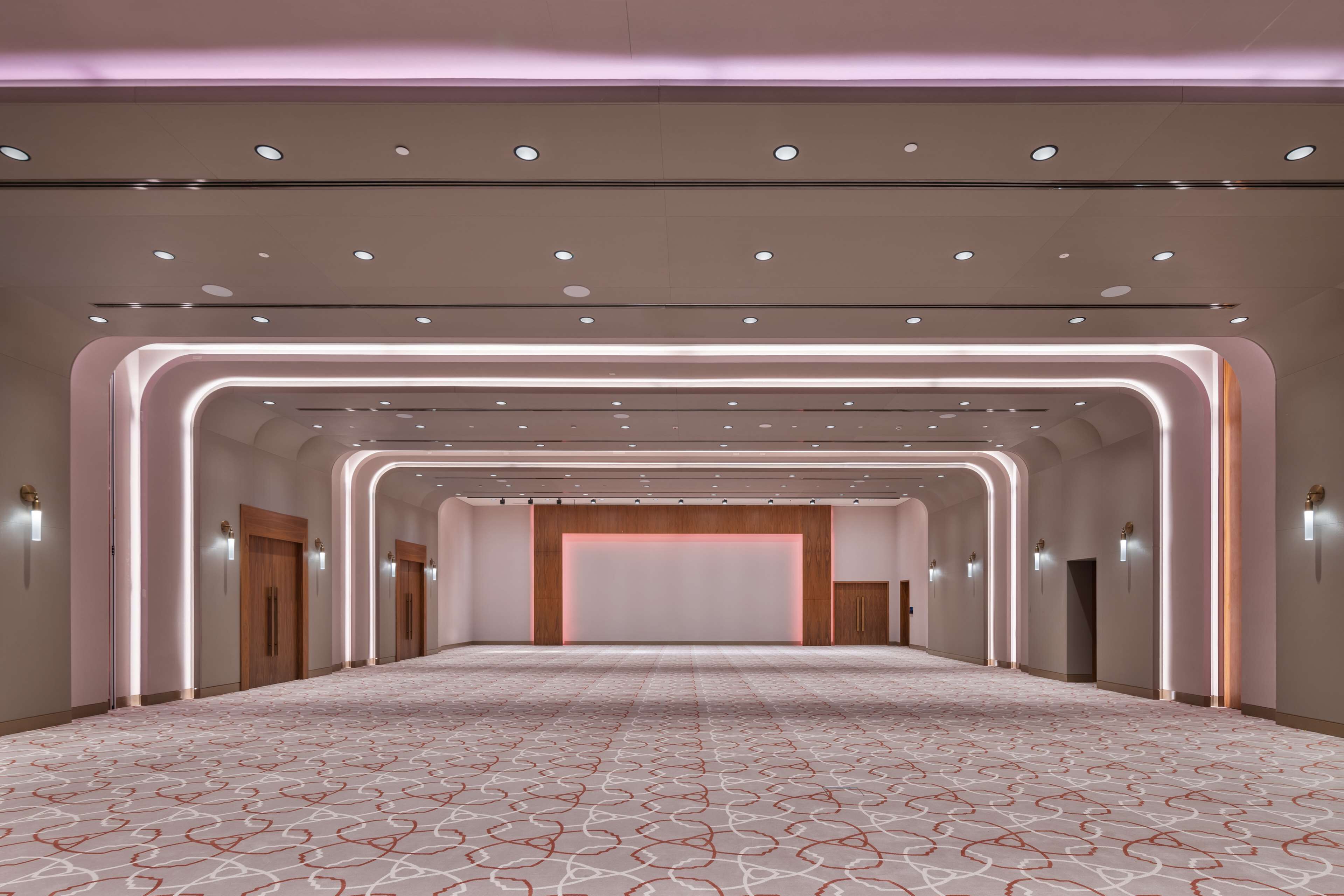 Un amplio vestíbulo de hotel con alfombra estampada, iluminación empotrada y una pared central con paneles de madera.