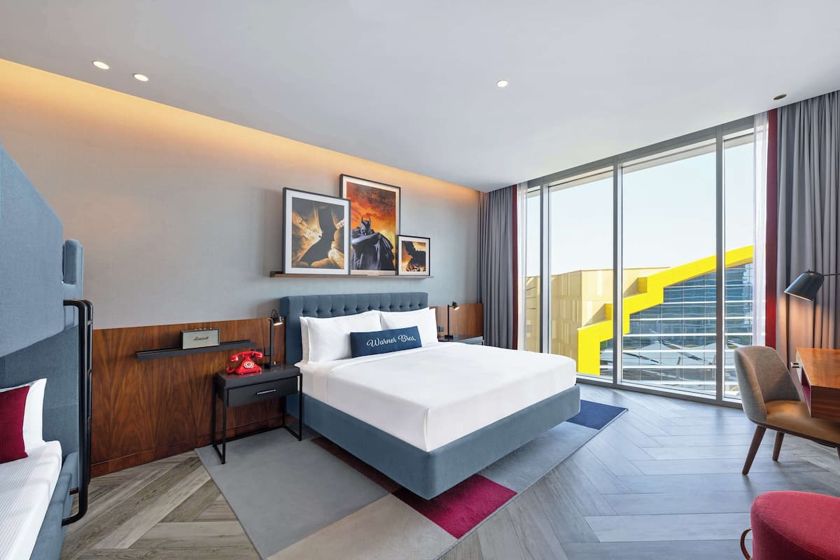 A modern hotel room with a large bed, a desk, a chair, and a view of a yellow staircase outside.