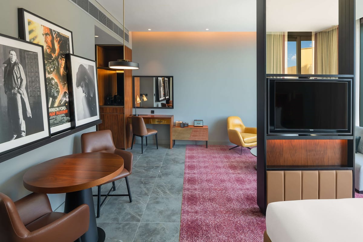 A hotel room with a bed, a TV, a desk, a chair, a table, and framed movie posters on the wall.