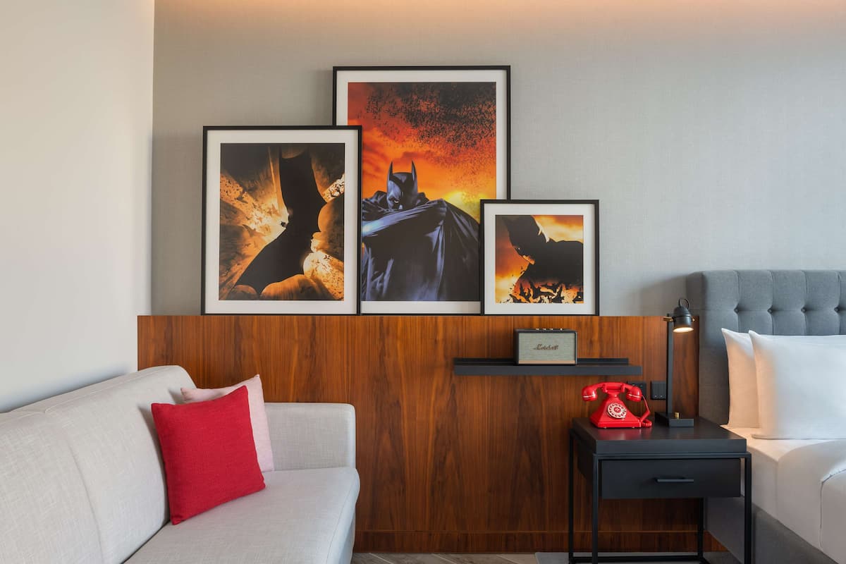 A hotel room with a wooden panel wall, a grey sofa with red and white cushions, a small bedside table with a red telephone, and framed artwork above the sofa.