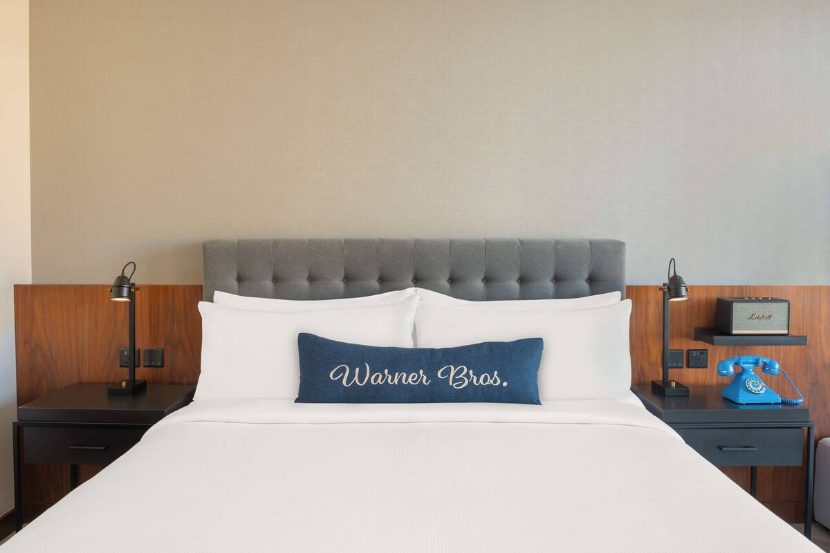 A hotel room with a tufted headboard, a bed with white linens, a blue pillow with "Warner Bros." text, a classic blue rotary phone, and a small shelf with a lamp and a picture frame.