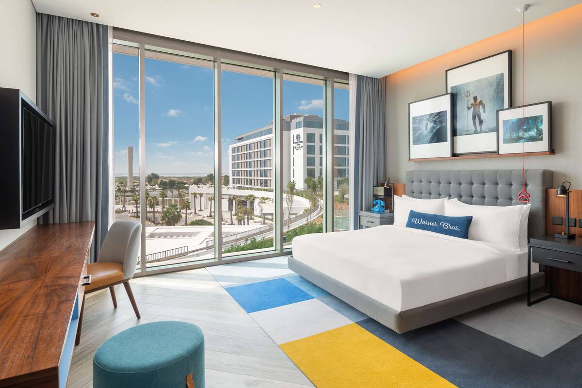 A modern hotel room with a large bed, a desk, and a view of a cityscape through floor-to-ceiling windows.