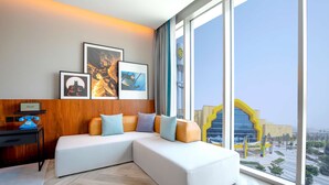 Una habitación de hotel moderna con un sofá modular, un escritorio y ventanas amplias que ofrecen vistas a un paisaje urbano y un edificio amarillo característico.
