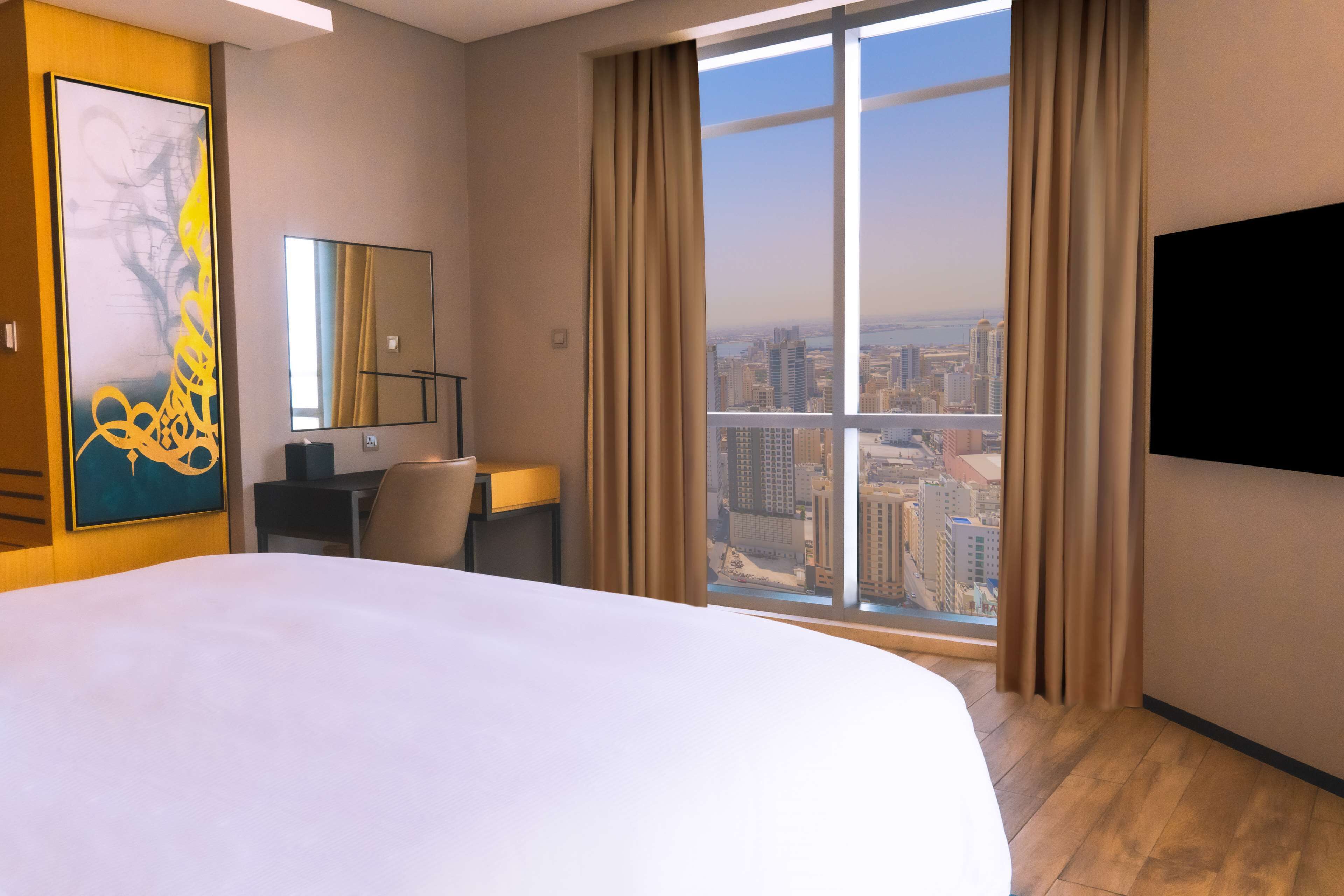 A hotel room with a large bed, a desk, a chair, a TV, a large window with curtains, and a city view.