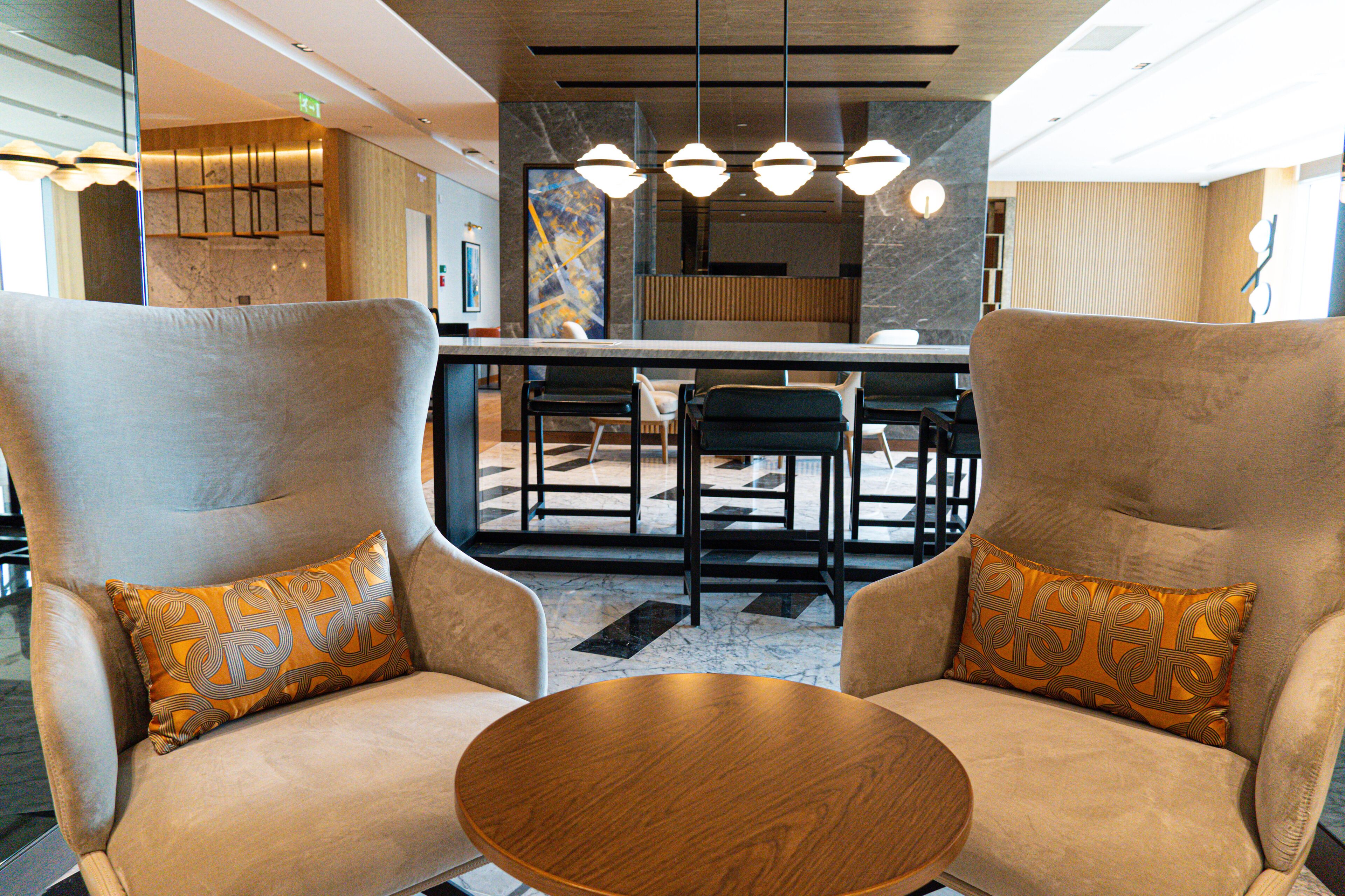 A modern hotel lobby with two armchairs, a round wooden table, and a bar area.