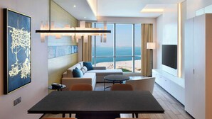 Une chambre d’hôtel moderne dotée d’un coin repas, d’un canapé avec des coussins, d’une télévision et d’une grande fenêtre offrant une vue sur la mer.
