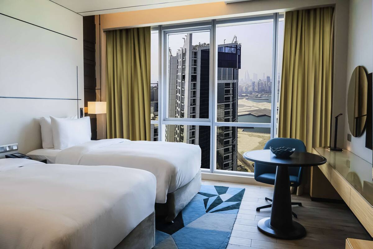A hotel room with two beds, a small table, a chair, a large window with curtains, and a view of the cityscape.