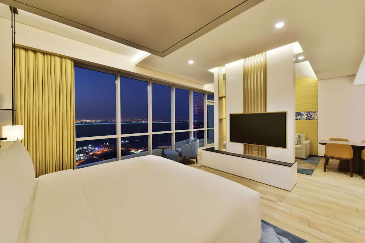 A hotel room with a large bed, a TV, a desk, and a view of the city at night.