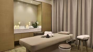 A modern spa room with a massage table, a mirror, a vanity with candles, and a large window with curtains.
